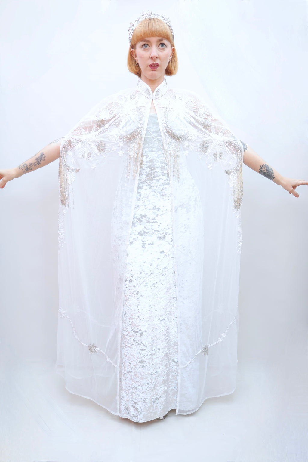 A model with short blonde hair and tattoos wears a white lace dress with a sheer white embroidered cape and a silver tiara, posing against a white studio background.