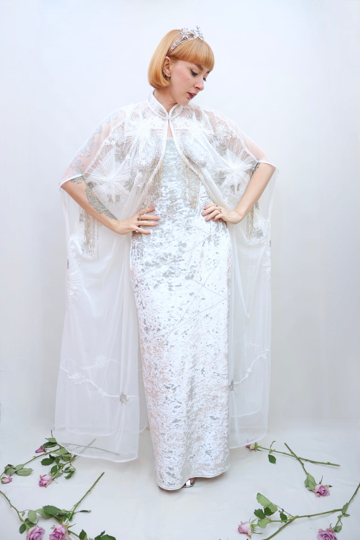 A woman modeling a white lace floor-length gown with a sheer white cape overlay, standing against a plain white studio background with scattered pink flowers on the floor.