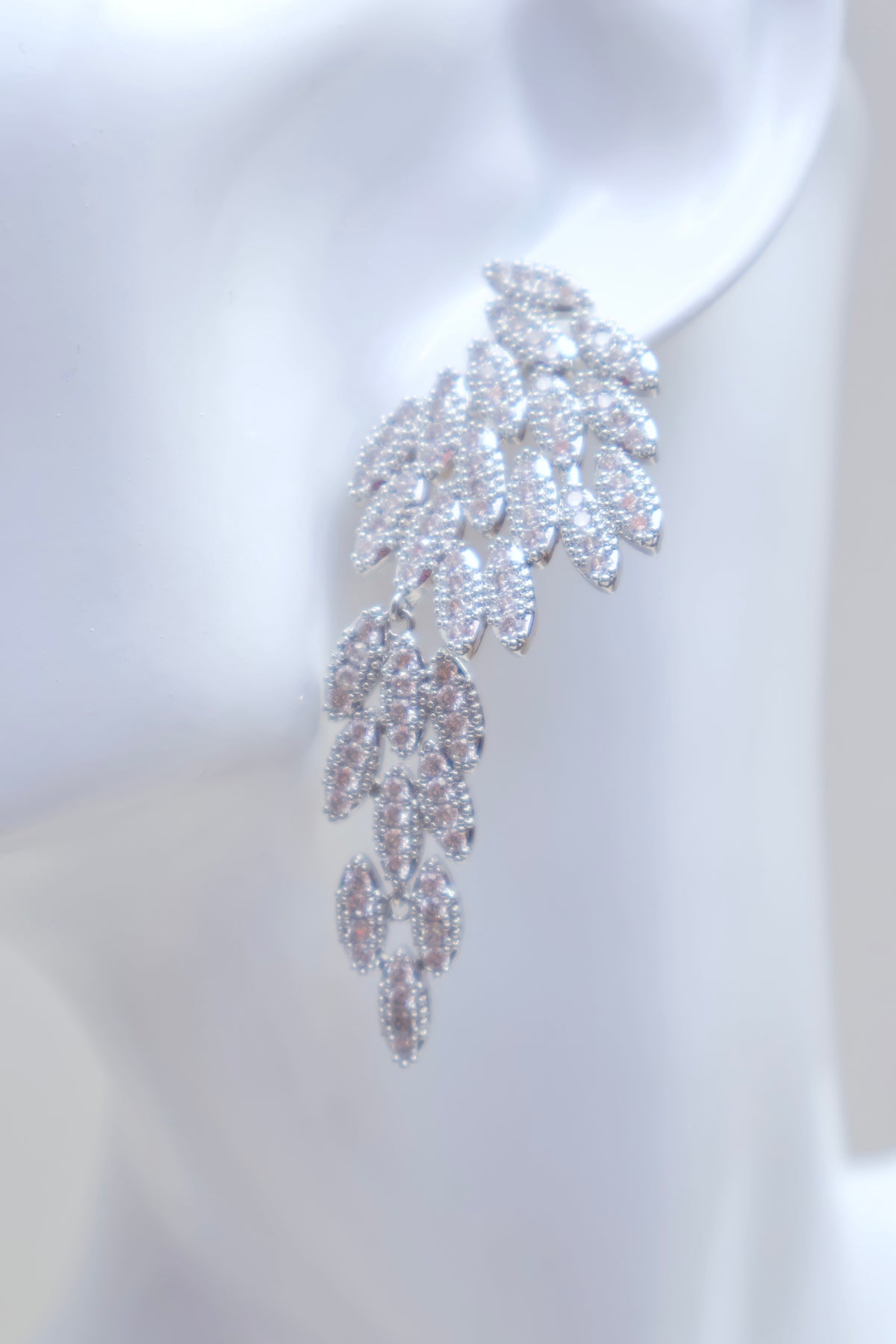 A single dramatic drop earring displayed on a white mannequin ear, featuring a silver-toned leafy vine design paved with clear stones.